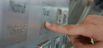 ein Finger drückt auf einen Aufzugknopf "Tür auf", der auch mit Braillezeichen beschriftet iste