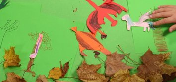 Von Kindern gebastelte Kulisse für einen Stop-Motion-Film: Buntes Papier und Laub als Hintergrund, ein ausgeschnittener Papierdrache wird von einer Kinderhand für die nächste Aufnahme bewegt.