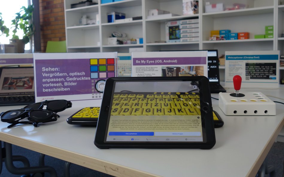 Station vo Erlebnisparcours: Mit der App Be My Eyes wurde eine sehbehindertenangepasste schwarz-gelbe Tastatur fotografiert und beschrieben. Im Hintergrund der Aussteller mit der Stationsbezeichnung Sehen: Vergrößern, optisch anpassen, Gedrucktes vorlesen, Bilder beschreiben. Daneben einige Sehbehinderungssimulationsbrillen