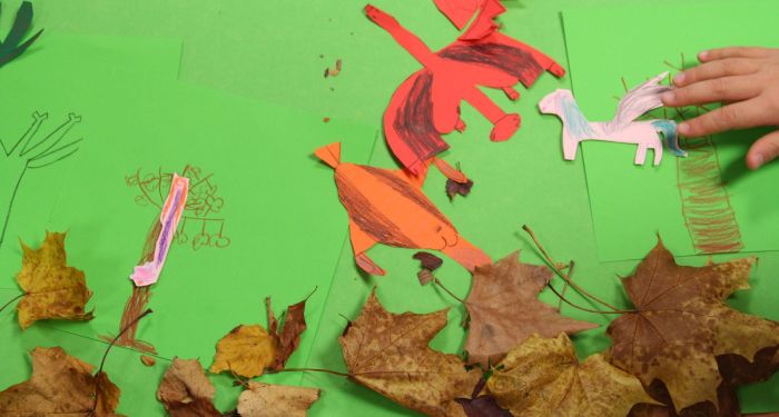 Von Kindern gebastelte Kulisse für einen Stop-Motion-Film: Buntes Papier und Laub als Hintergrund, ein ausgeschnittener Papierdrache wird von einer Kinderhand für die nächste Aufnahme bewegt.