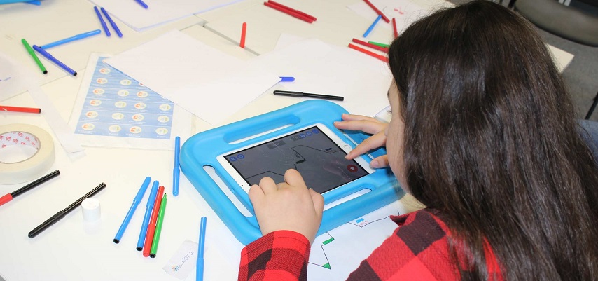 Mädchen mit Tablet und der App Draw Your Game. Vor ihr weiße Blätter und bunte Stifte, mit der die Level gezeichnet werden.